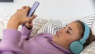 Teenage girl lying down wearing headphones and reading her phone.