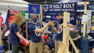 A group of students wearing the same "12 Volt Bolt" team t-shirts stand in front of their workspace