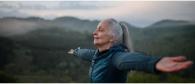 Person opens arms to sky with eyes closed with mountains behind them