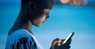 Close-up of a young woman using a smartphone at dusk, her face illuminated by the screen’s glow. She appears focused, with a soft blue background and blurred city lights in the distance.