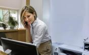 A woman staring at a computer screen while on the phone. The woman is smiling and is in an office setting.