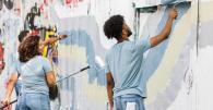 Group of young adults painting over a graffiti-covered wall with rollers and brushes, working together as part of a community mural or cleanup project. They wear casual clothes and appear engaged and cheerful.