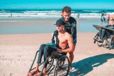 A person in a wet suit helping another in a wheelchair on a beach.