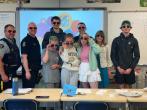 A group of adults and kids all wearing rainbow-rimmed glasses in a classroom setting. Some adults in police uniform.