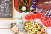 Party materials on a table for Super Bowl Sunday: Football, hard boiled eggs decorated like footballs, wings and a small chalk board with a play written in chalk.