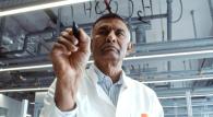 Dr. Ponnusamy wears a lab coat writing out chemical compounds on glass with a marker.
