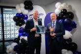 Two men standing in front of balloons. One is holding a framed certificate and one is holding an award.