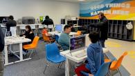 A group of children sitting at desks with computers. A row of cabinets for coats and bags on the side wall under a mural "Good Things Happen Here."