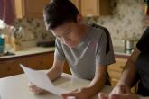 Young boy reading from a piece of paper at a kitchen table.