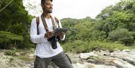A smiling person with a camera and notepad next to a stream