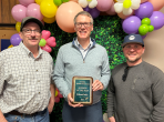 Three smiling people posed with a plaque. Balloons behind them.