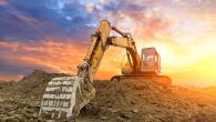 A large construction digger on a hill of dirt, a bright setting sun behind it.