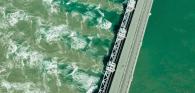 Aerial view of a large dam with green water on one side and cloudy green turbulent water on the other.