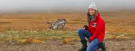 A person crouched on an open plain, a caribou in the distance