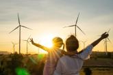 an adult holds a child and each have an arm stretched out. The sun is setting behind a filed of wind turbines