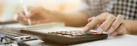Close up of a person using a calculator, writing with the other hand and a laptop in front of them.