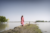 A woman stands on a narrow embankment surrounded by floodwater, with trees and people wading in the distance.