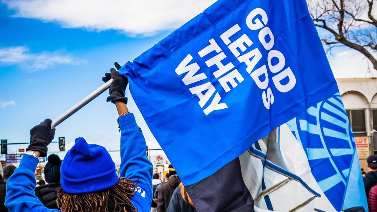 Person wearing blue holds a blue flag with the words, "good leads the way" printed in white on the flag