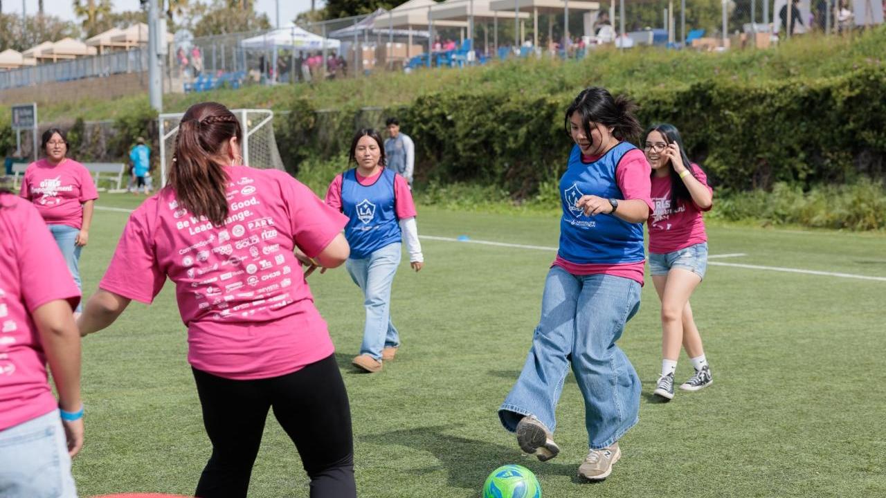 Girls wearing pink Girls Empowerment Day shirts play soccer during a youth sports clinic on an outdoor field.