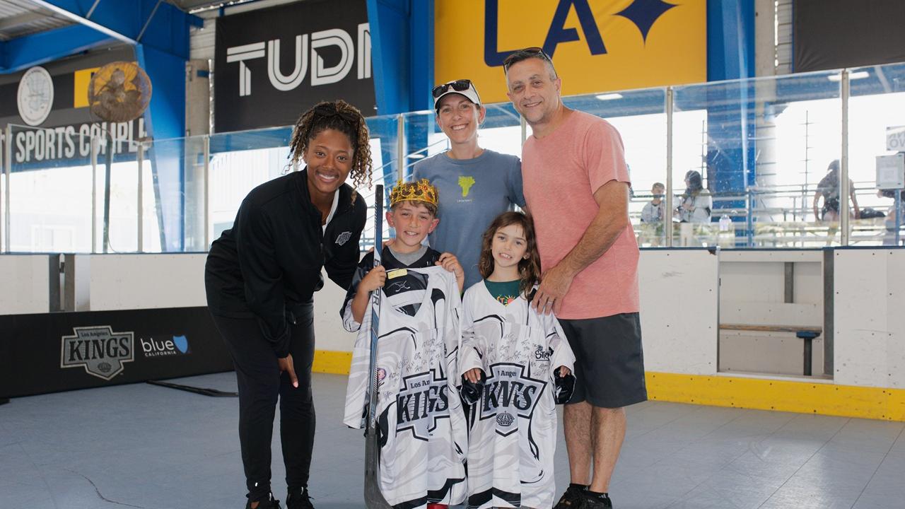 Two youth participants stand with signed LA Kings jerseys alongside family members and LA Kings Blake Bolden during a ball hockey event at an indoor sports facility.