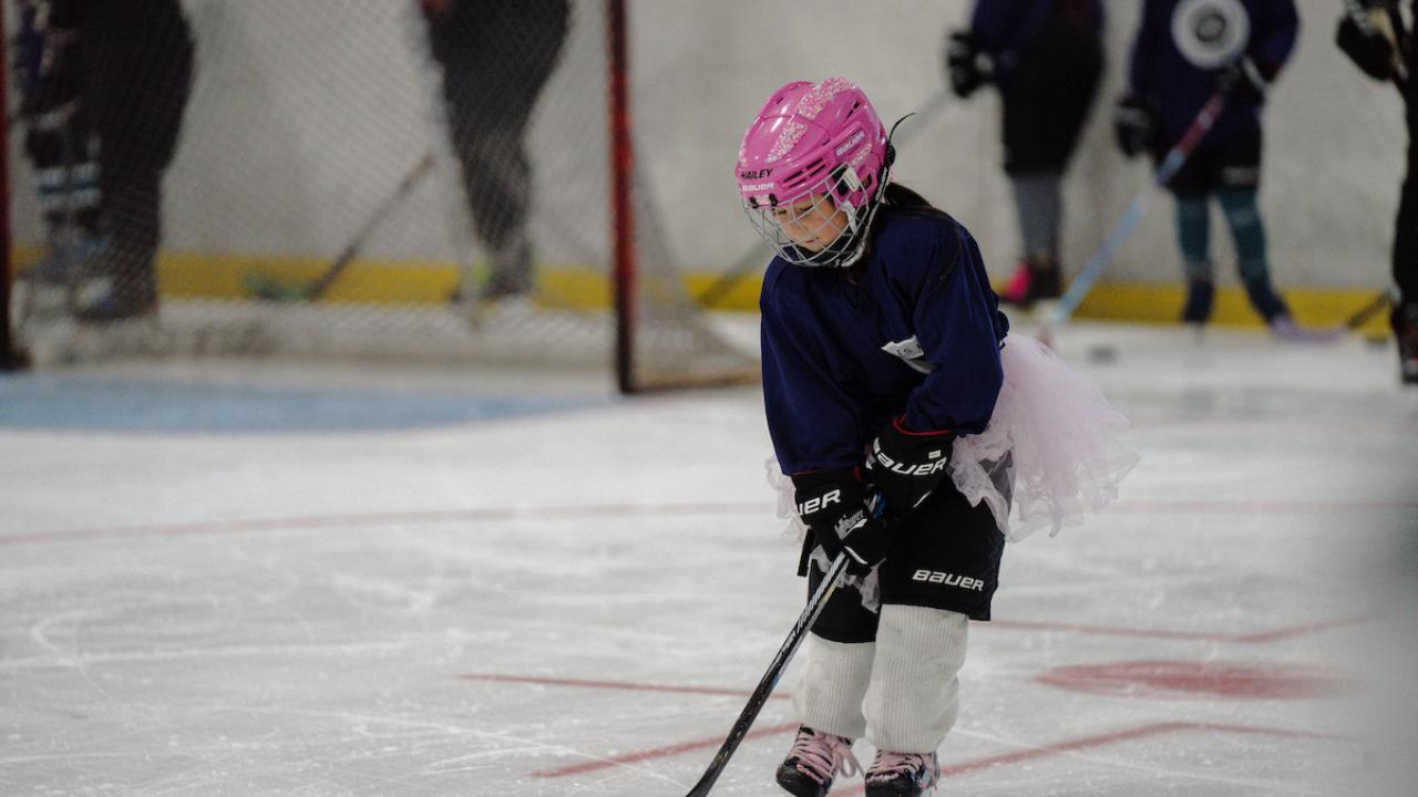 Participant in full hockey gear skates on the ice while practicing puck control during LA Kings Girls Camp. A pink helmet and matching hockey stick stand out against the rink background.