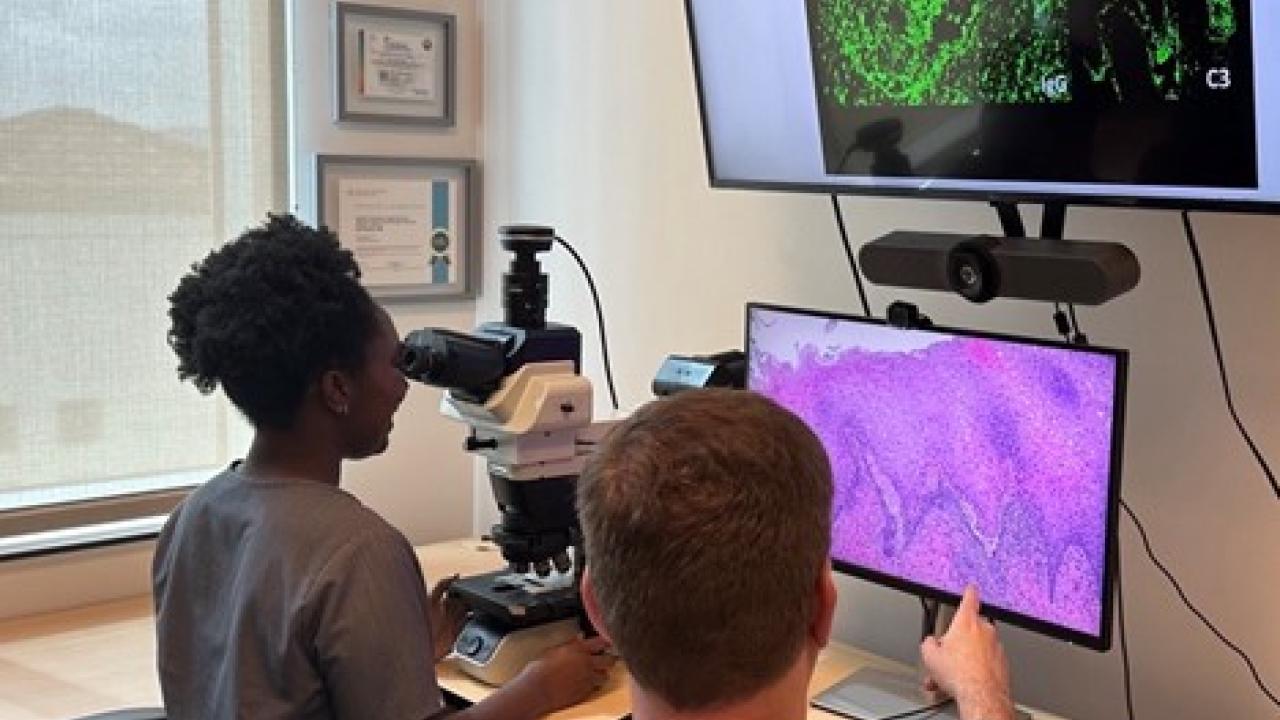 A person looks through a microscope while a person in a white coat points to a screen