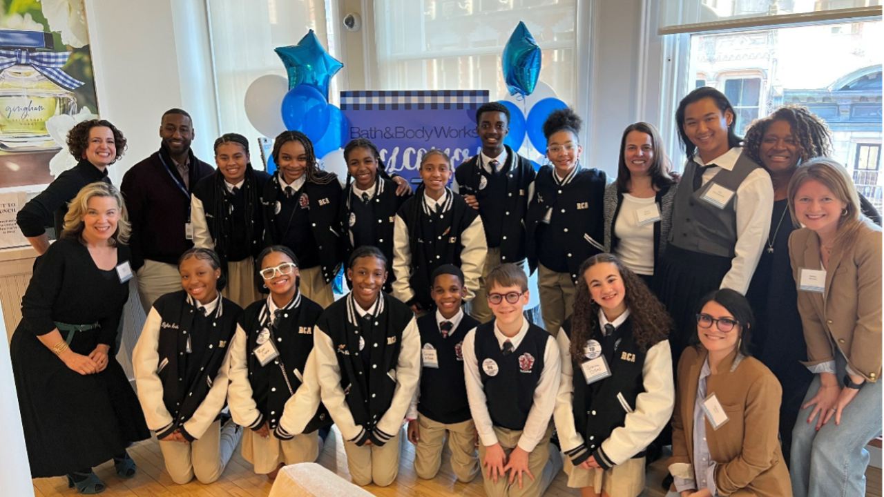 12 middle school students smile alongside seven Bath & Body Works employees in front of a sign with balloons and sunny windows.