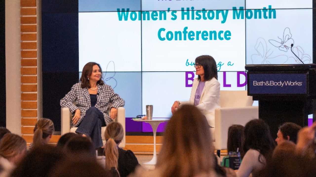 Two women sit together onstage in front of a crowd of employees with a screen behind them.
