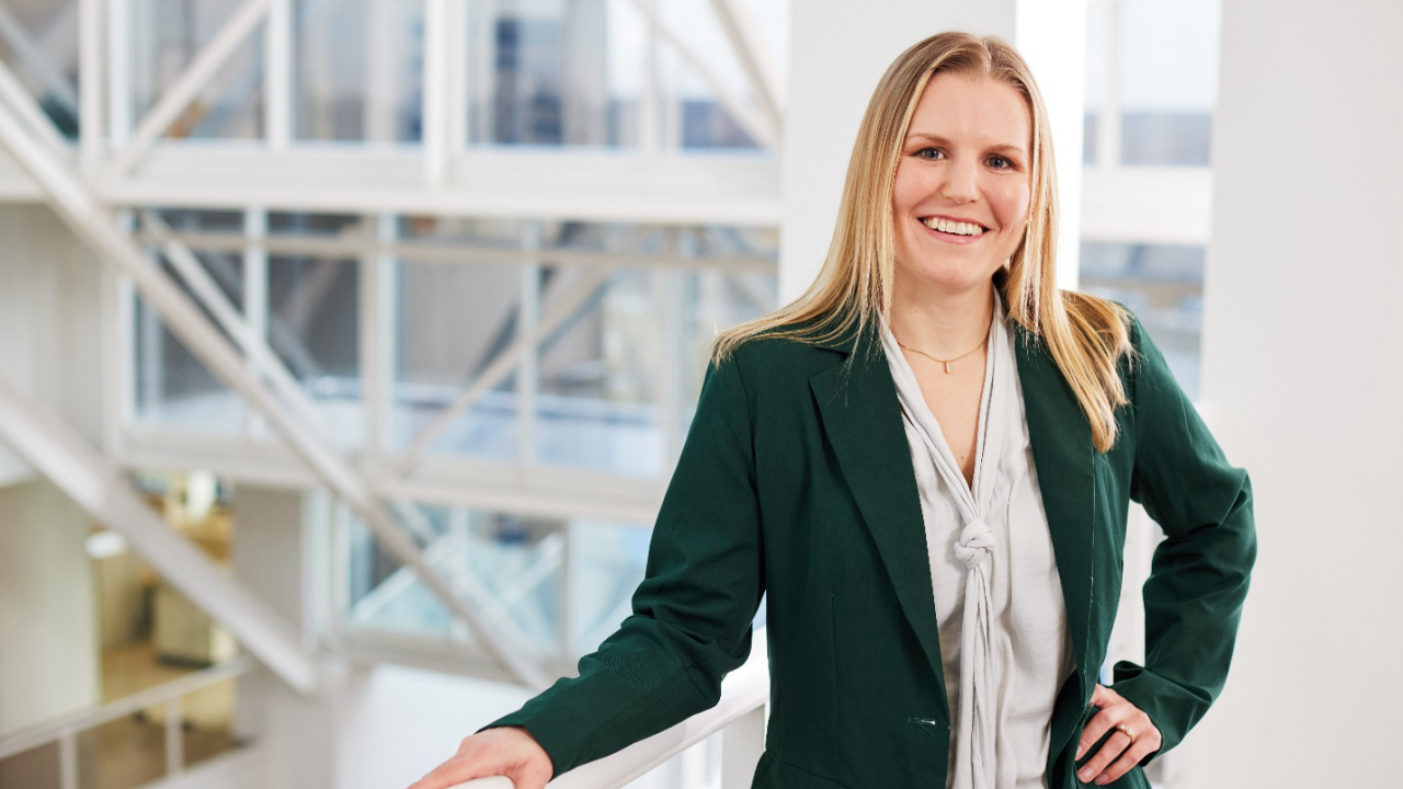 Maureen McHugh smiles at the camera and stands holding a railing at the top of a set of white steps at Bath & Body Works’ home office in Columbus, Ohio. Sun shines through a wall of windows in the background.