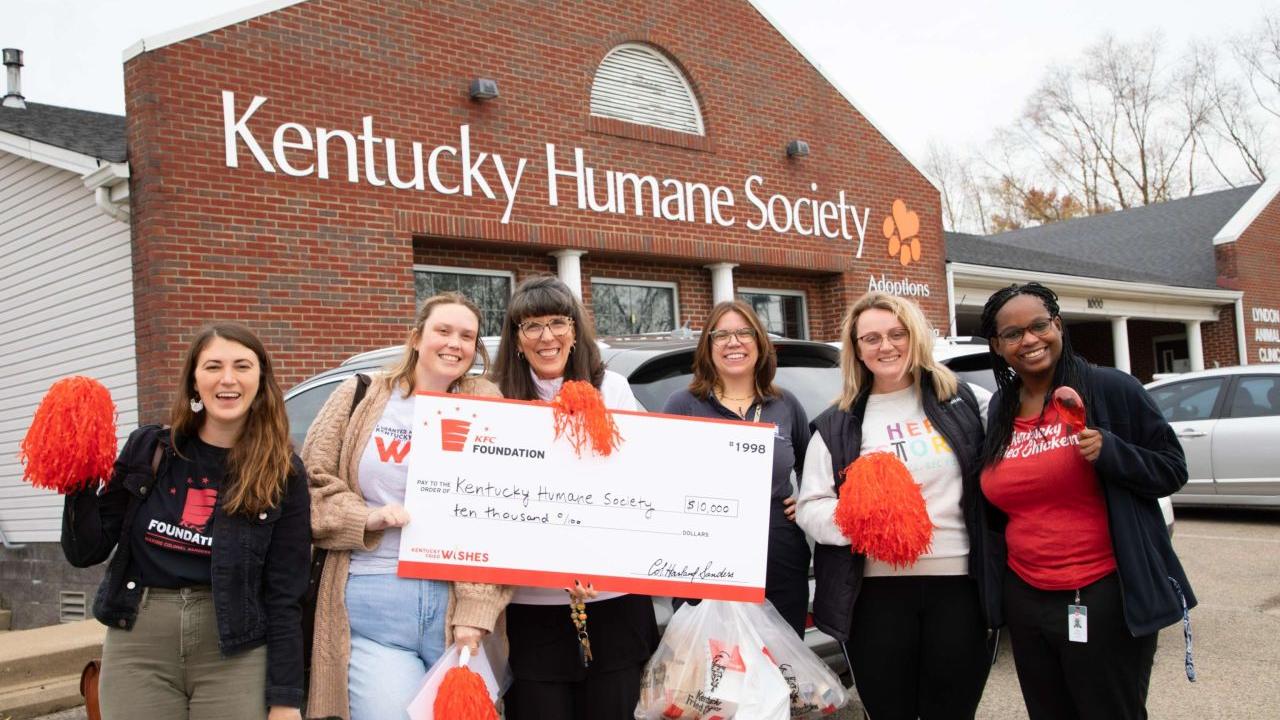 Six people standing together outside in front of the Kentucky Human Society, holding a large check