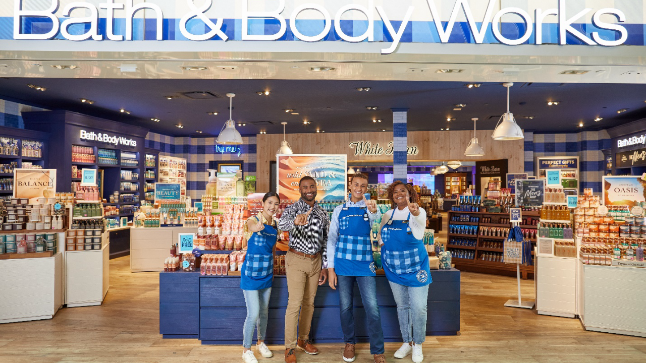 Four Bath & Body Works associates stand at the entrance of a store smiling and giving a “number 1” hand signal to the camera. A colorful selection of candles, hand soaps, lotions and other products are displayed on tables behind them.