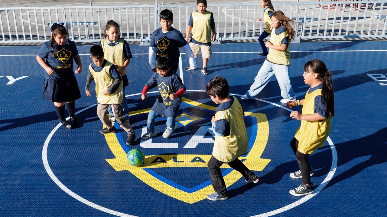 Children wearing LA Galaxy jerseys and pinnies play soccer around a ball on a branded mini-pitch court.