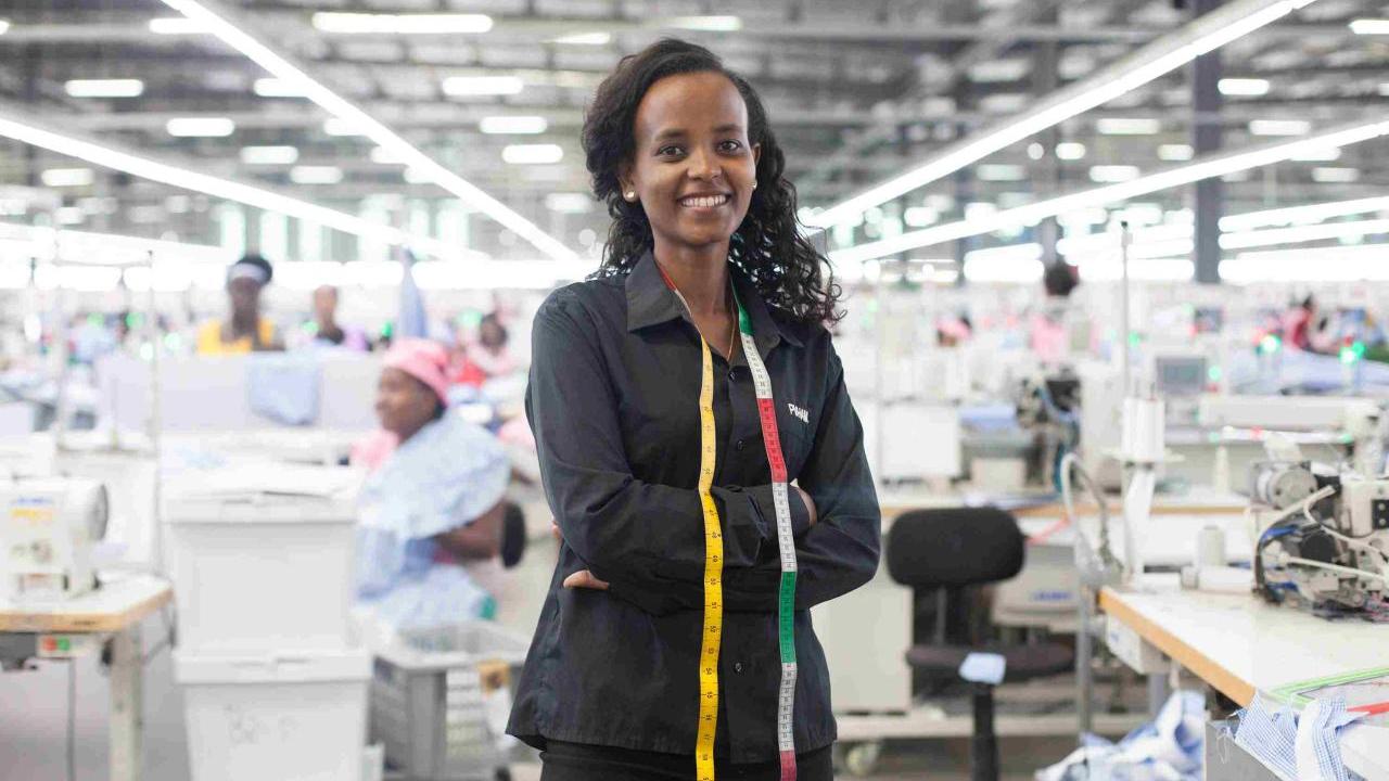 A young women in black clothing stands in front of a large room of women working on sewing machines. 