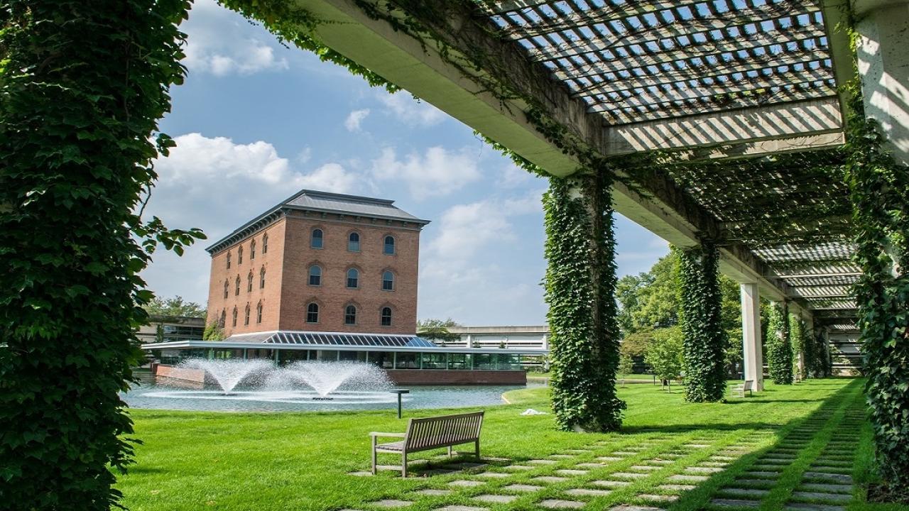 From under a plant-covered pergola, A rectangular brick building behind a water feature.