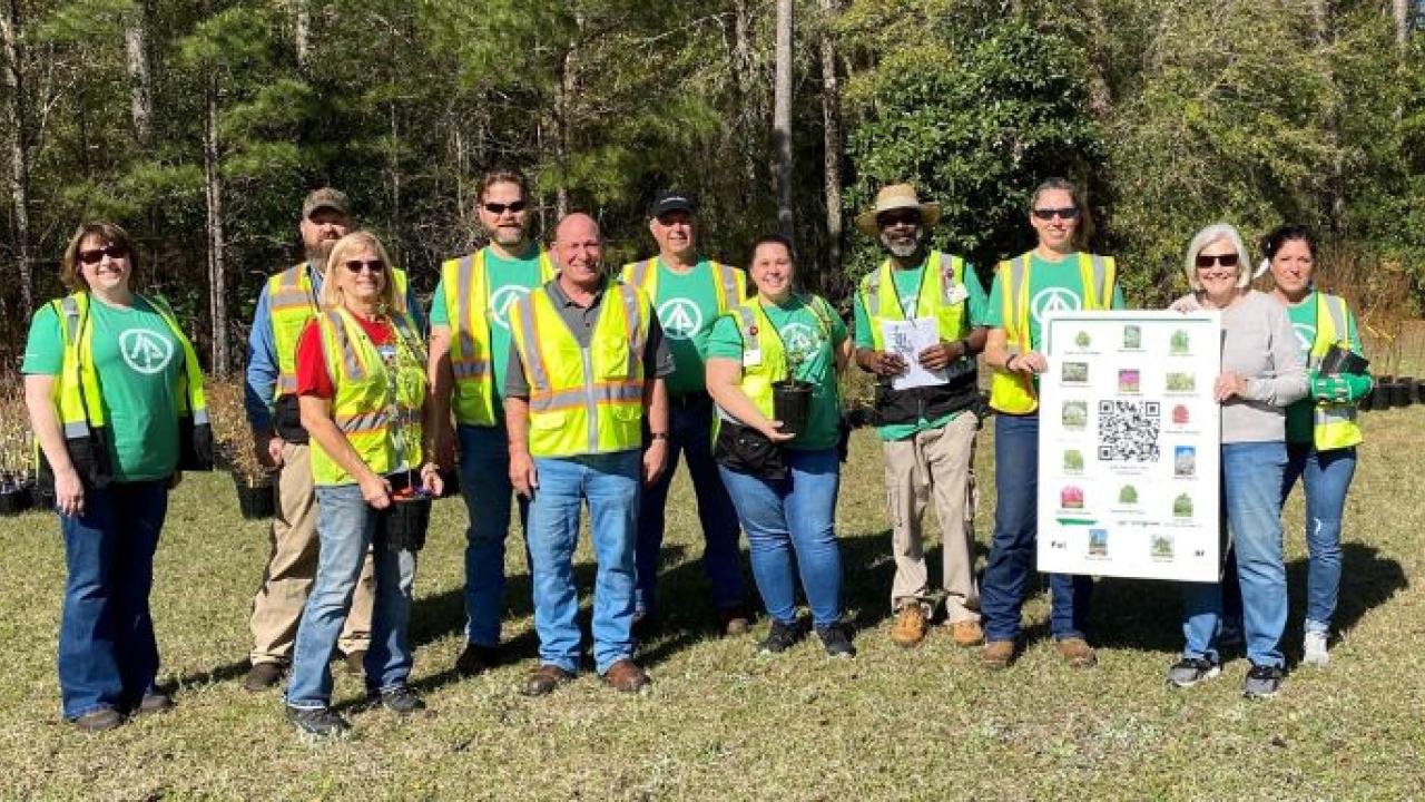 International Paper volunteers in safety vests holding potted trees and a poster of the kinds of trees planted