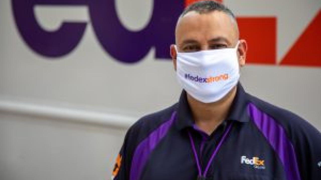 Employee in uniform stands in front of FedEx truck, wearing a mask that says #fedex strong