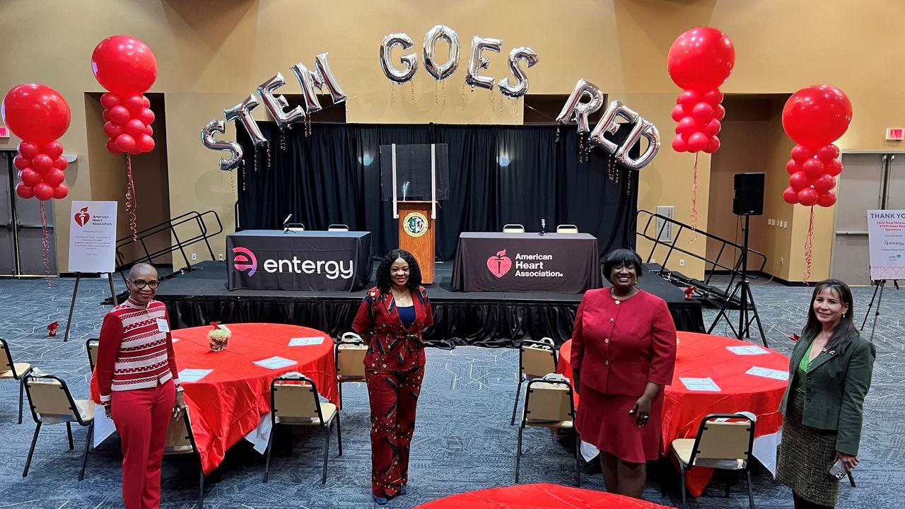Four women standing in front of a stage featuring balloons that read "STEM GOES RED"