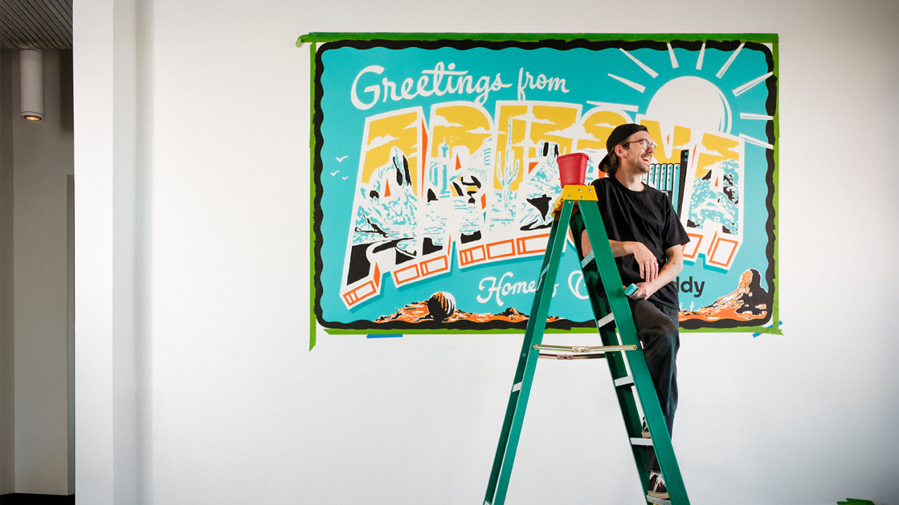 Artist on ladder in front of mural which reads "Greetings from Arizona"