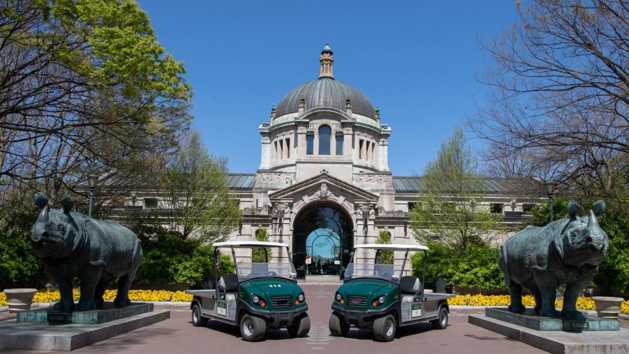 Two electric utility vehicles are parked in front of Bronx Zoo's Central Building on a sunny day. Flanking the vehicles are two animal statues.