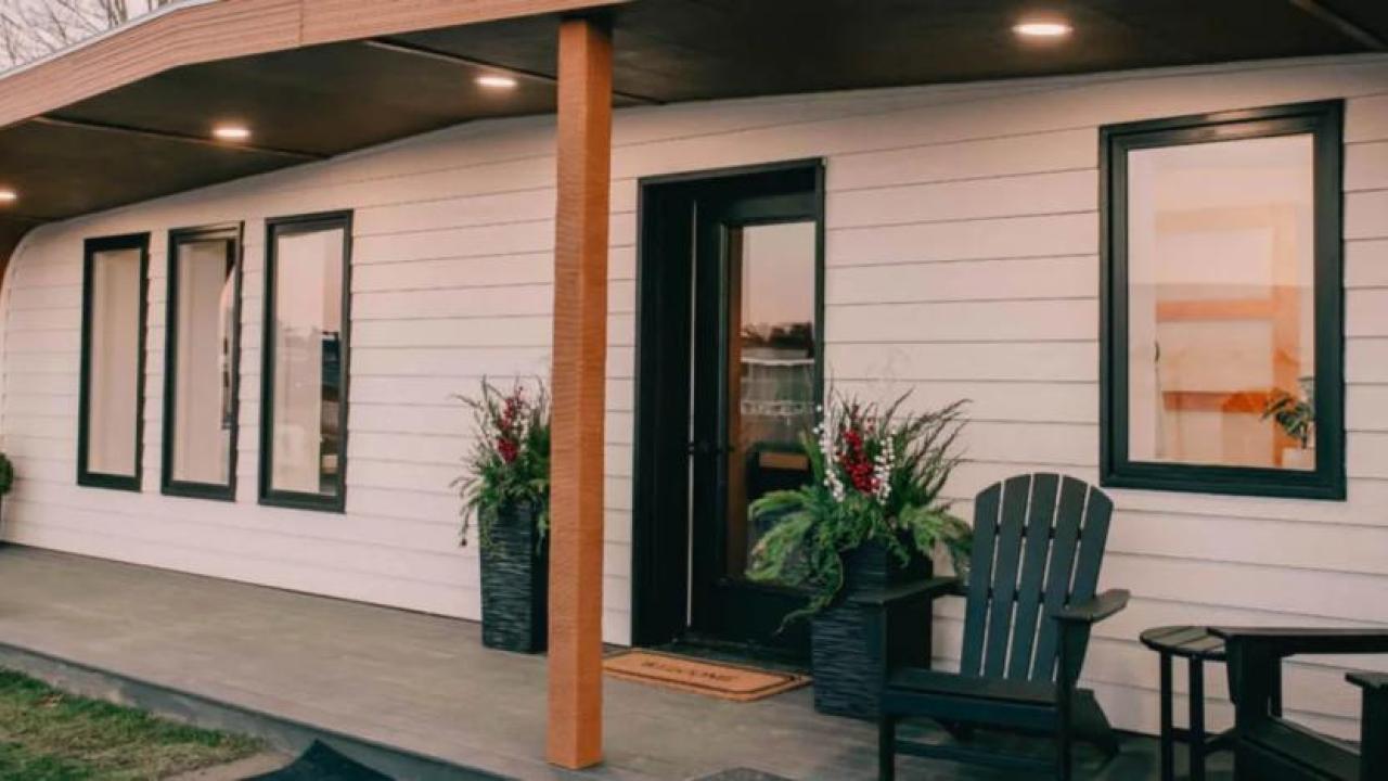 Exterior of the 3D printed home's front porch. Two chairs and a table to the side, plants on the sides of the doorway.