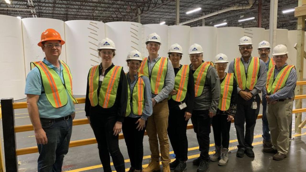 A group of people posed in a large warehouse. Each wearing a hard hat and high-vis vest.