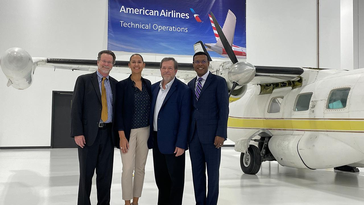 Four people standing in front of a small plane in a hanger, American Airlines banner on the wall behind them