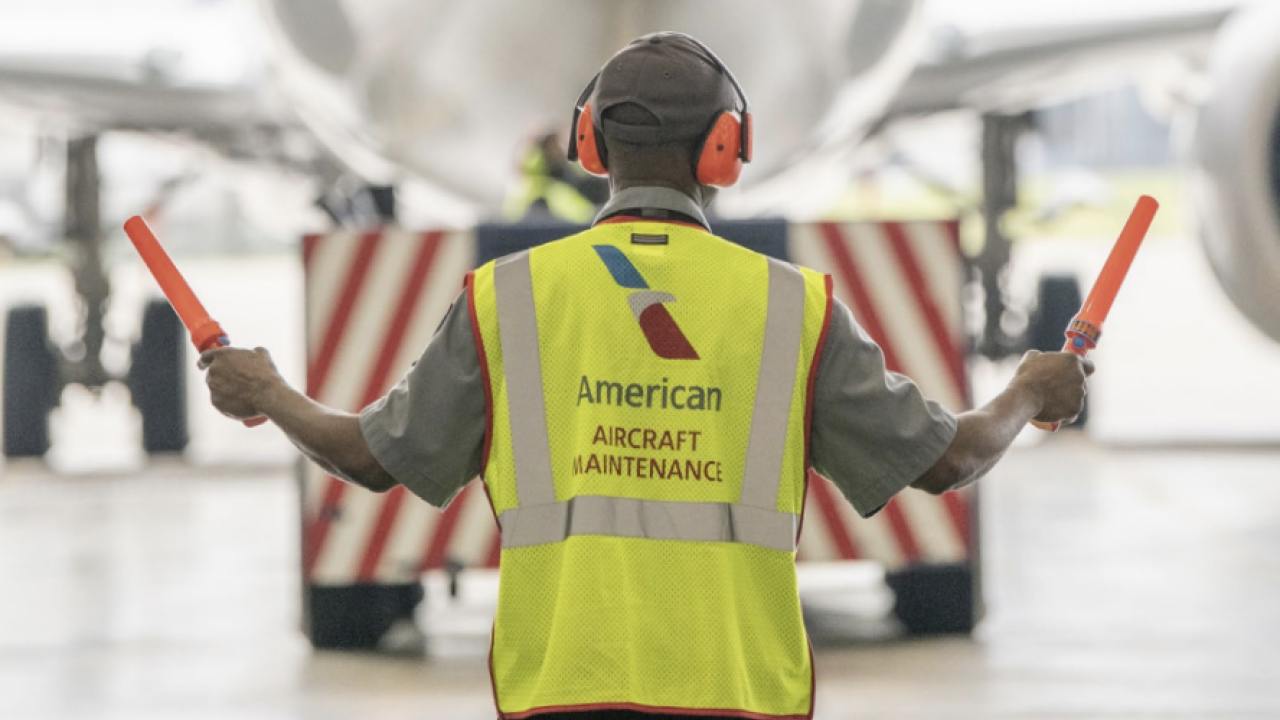 A person guiding a plane wearing a hi-vis vest that reads "American aircraft maintenance" 