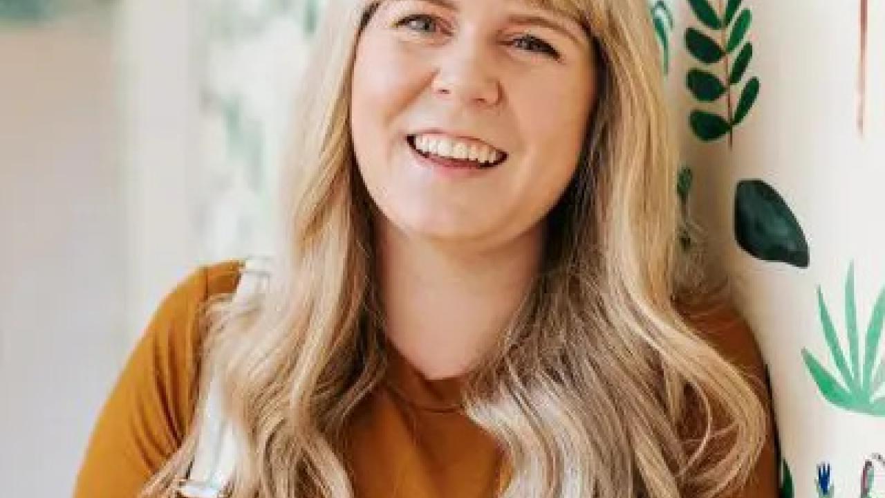 Ally Watson, smiling, leaning on a wall with painted plants.