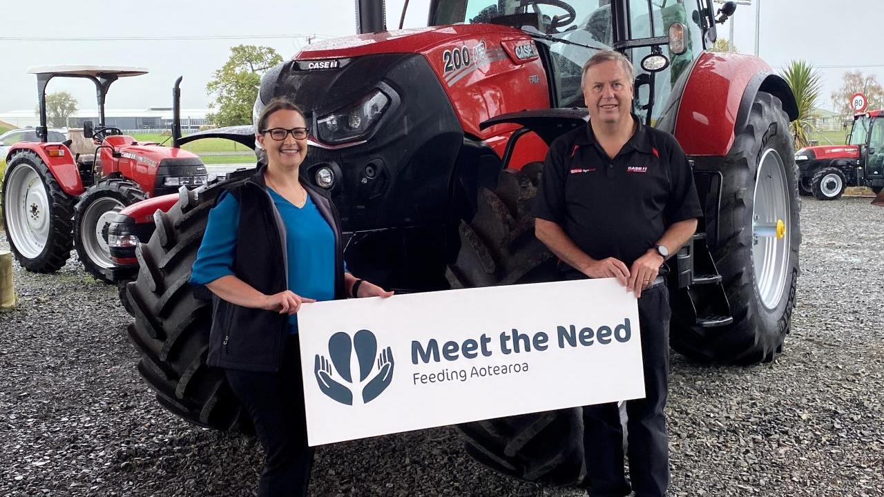 Two people stand in front of a tractor with a "Meet the Need" sign