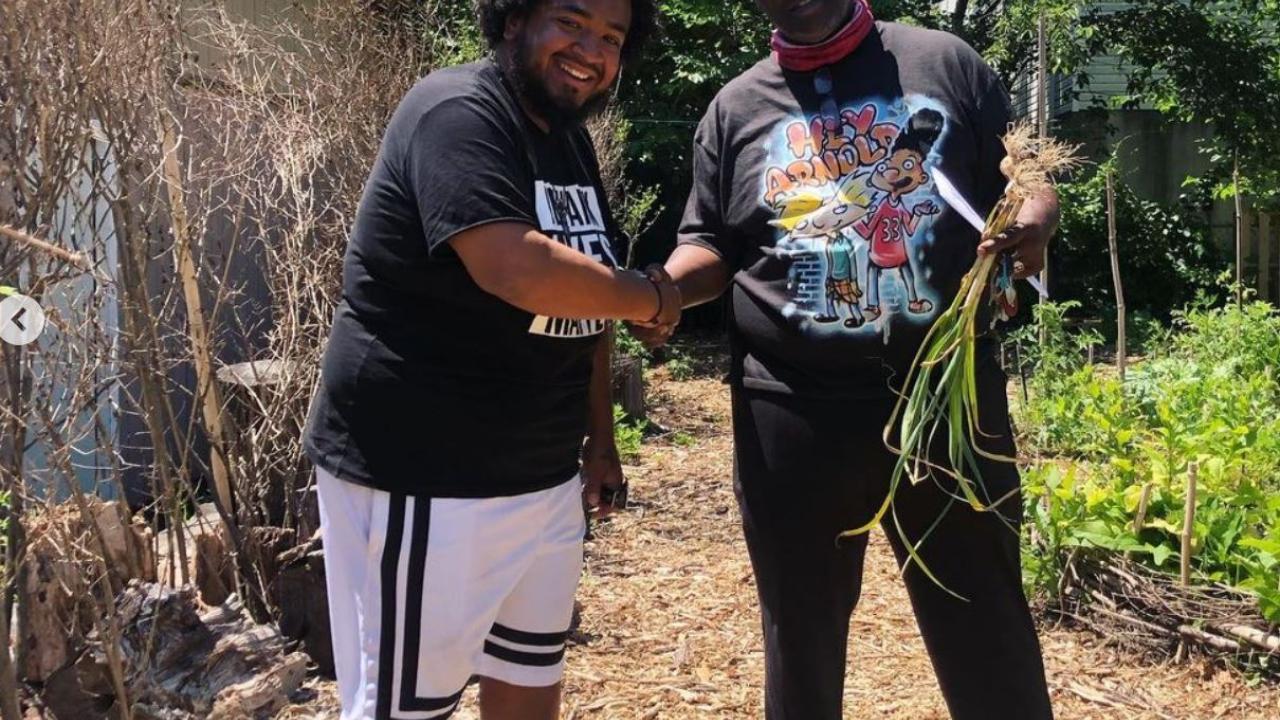Two people shaking hands while looking at the camera. One holds a bundle of produce. They're standing on a mulch path in a vegetable garden.