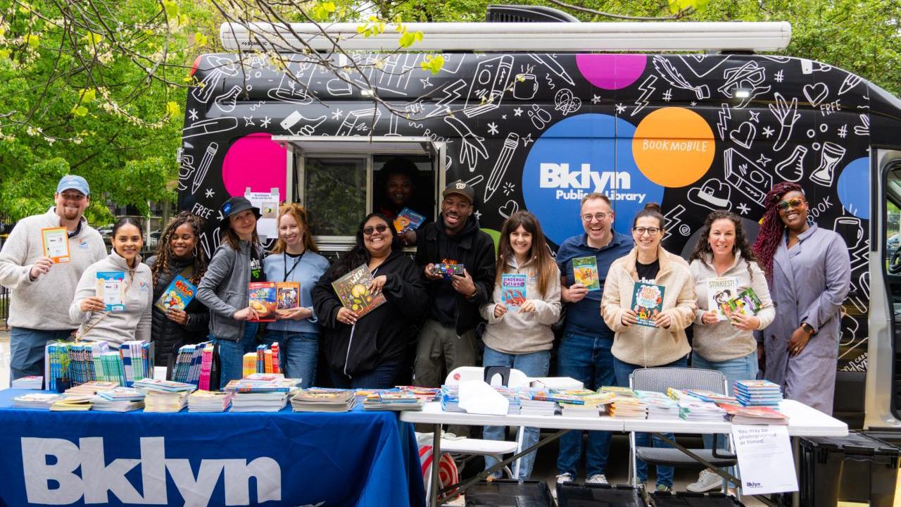 A group of people holding books, stood behind a table that has a Brooklyn Public Library banner draped over 