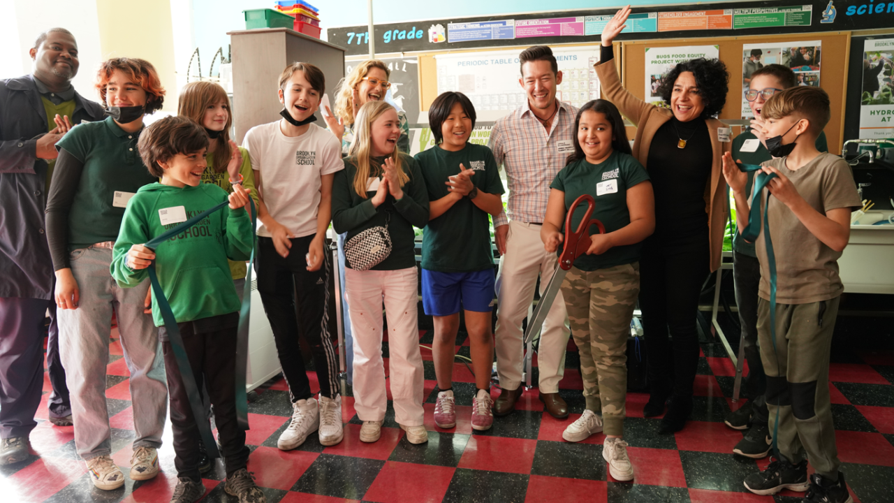 A group of adults and children are standing in a classroom clapping as a student cuts ribbon with oversized scissors. 