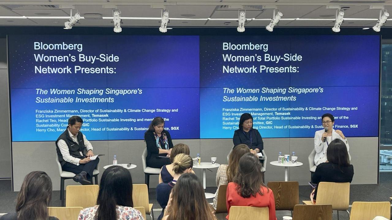 Four people on a stage, a small white table next to each of them. A small audience in front of them. Two digital projection screens behind them displaying "Bloomberg Women's Buy-Side Network Presents: The Women Shpaing Singapore's Sustainable Investments."