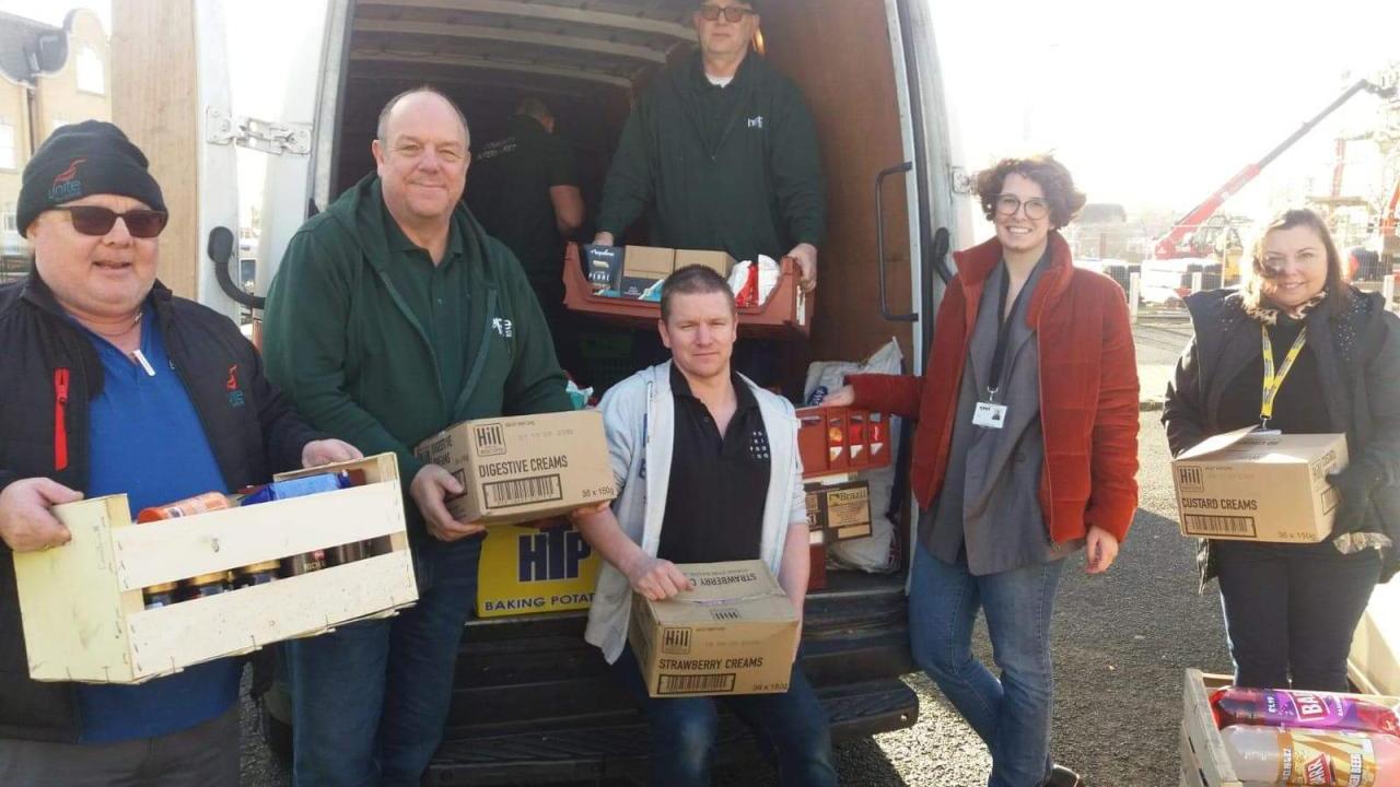 A group of six people carrying boxes, smiling at the camera. An open truck behind them with a person inside.